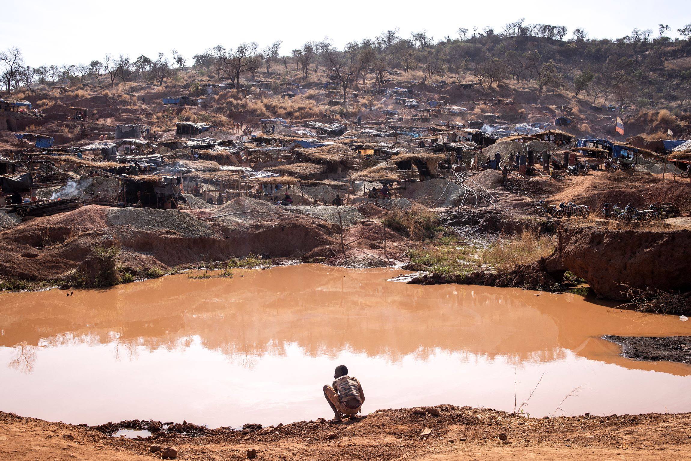 A man crouching beside a large open pit filled with water and surrounded by huts. 